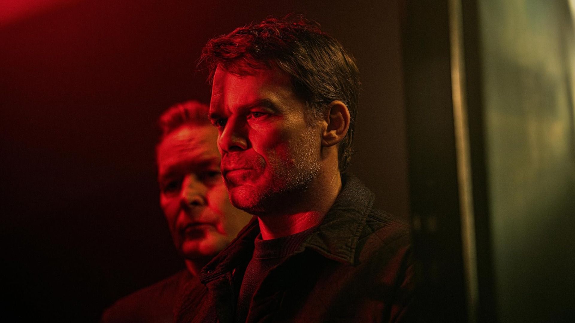 Actor Michael C. Hall stands in profile in red lighting with part of a man's face visible behind him