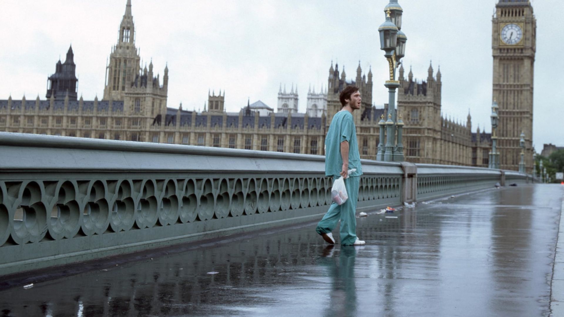 Actor Cillian Murphy in blue clothing stands on an empty bridge with London's Big Ben clock tower visible nearby