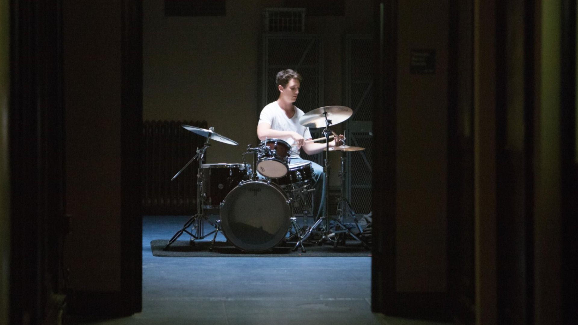 Dimmed lighting, actor Miles Teller plays drums in a white t-shirt