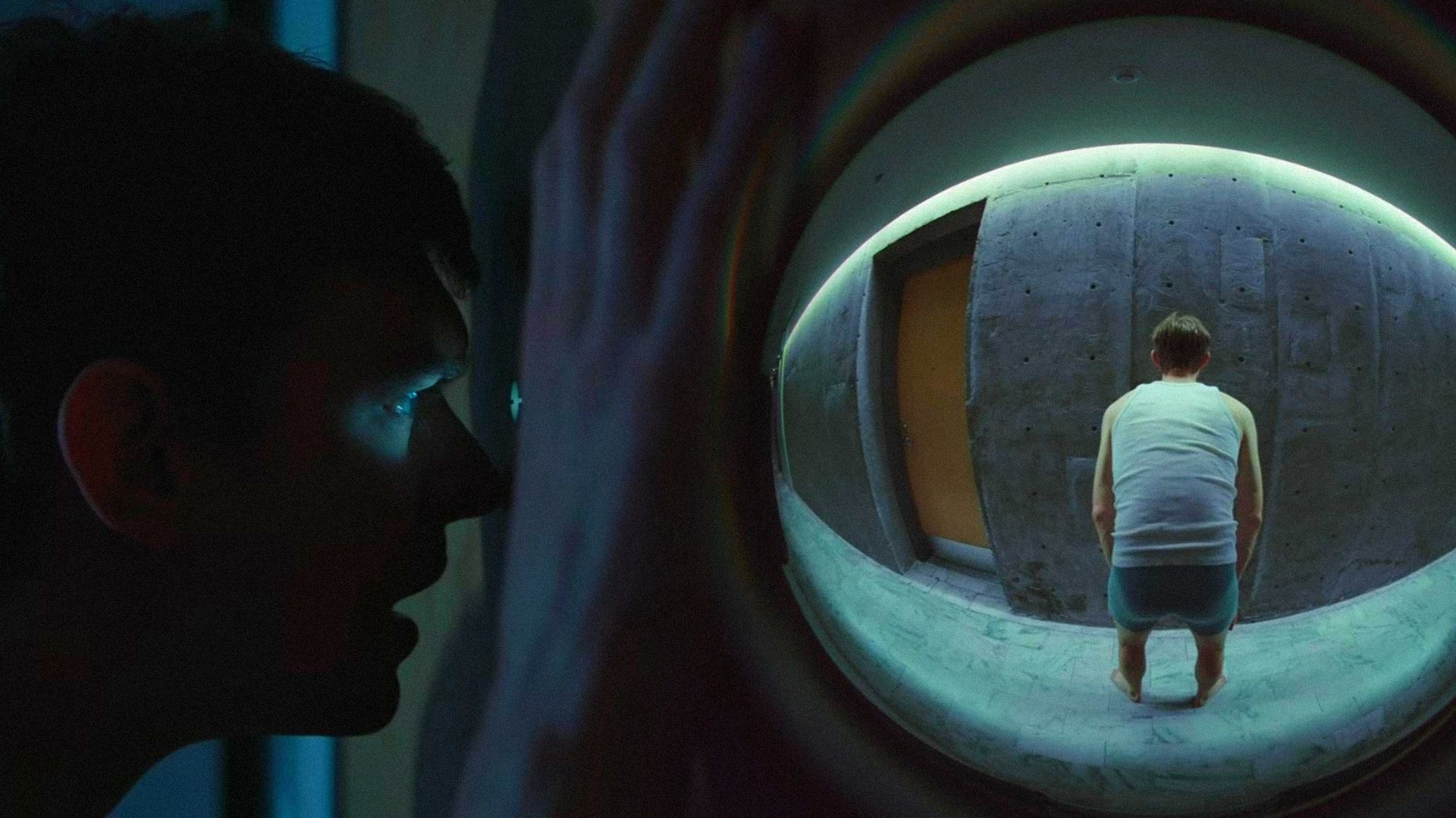 A man looks through the door peephole, behind the door stands a man in a white tank top and underwear