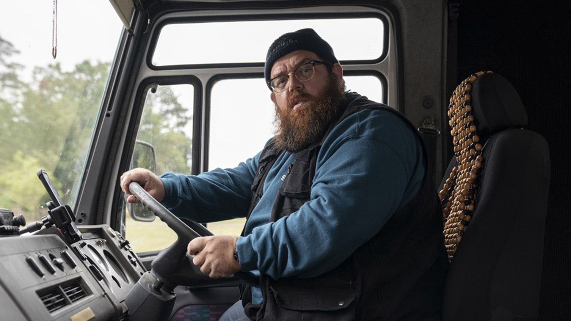 Nick Frost wearing glasses and a hat sits behind the wheel of a truck, looking at the camera