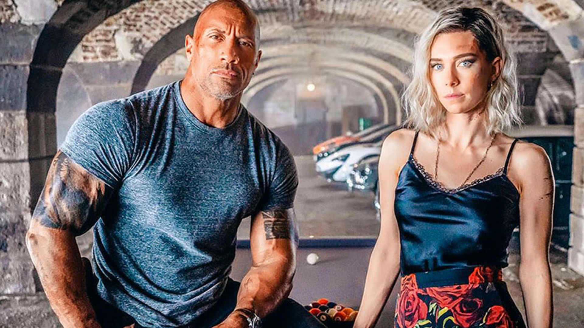 Dwayne Johnson in a tight-fitting t-shirt and Vanessa Kirby in a tank top and patterned skirt pose against a backdrop of arches and sports cars