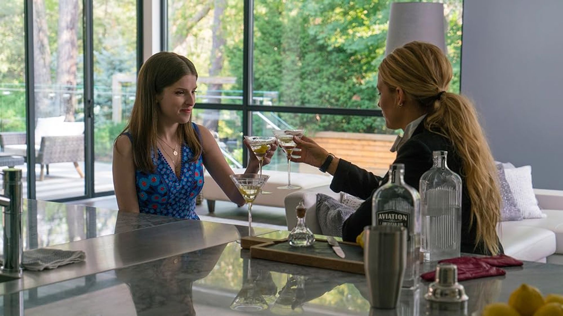 Anna Kendrick and Blake Lively clinking wine glasses while sitting in the kitchen