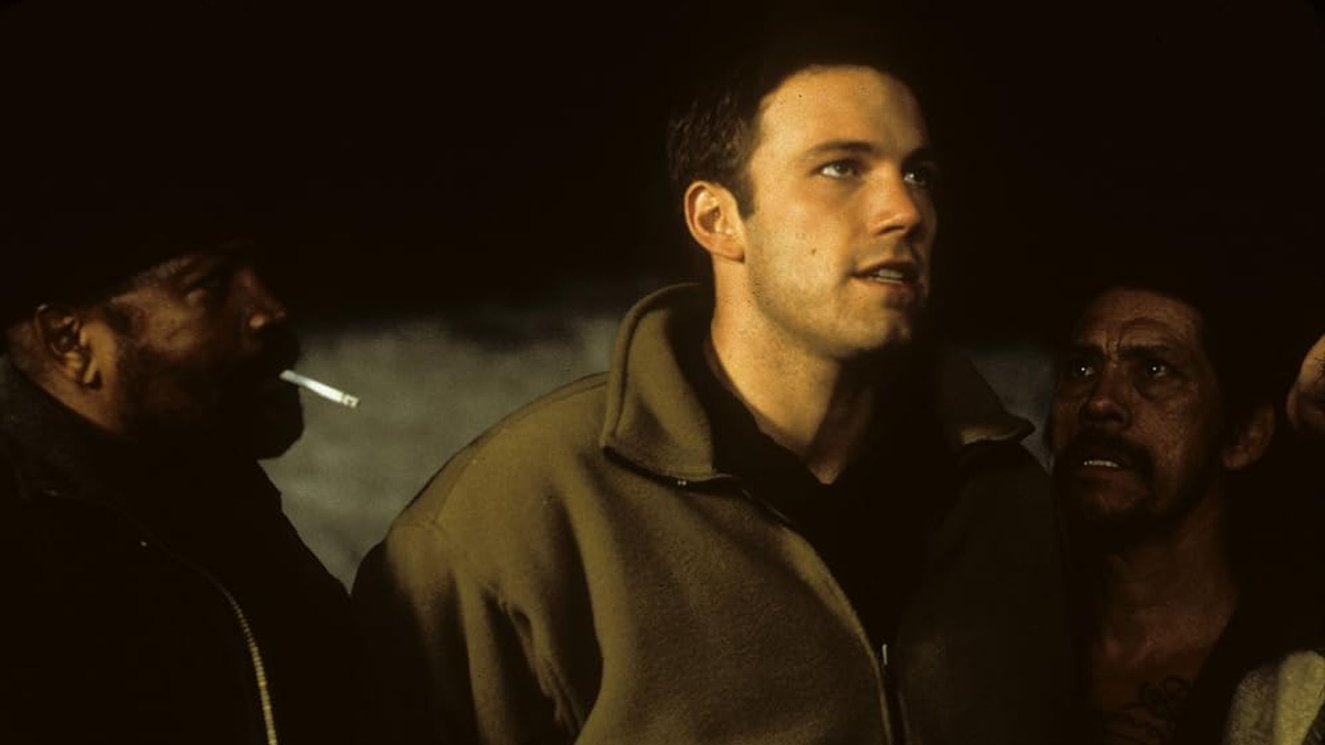 Ben Affleck in a beige jacket stands between two people, one of whom is smoking, in a dimly lit setting