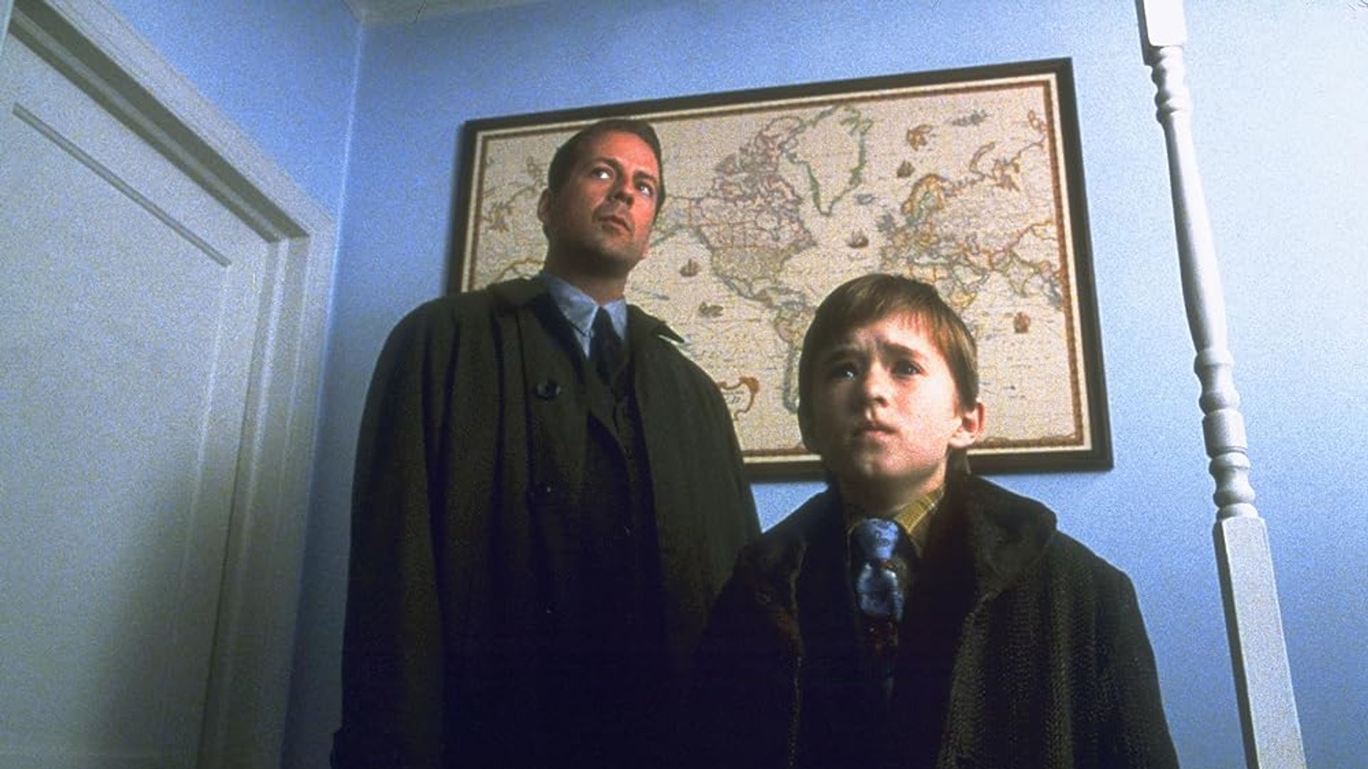 Bruce Willis and Haley Joel Osment standing in a hallway with a world map hanging on the wall behind them