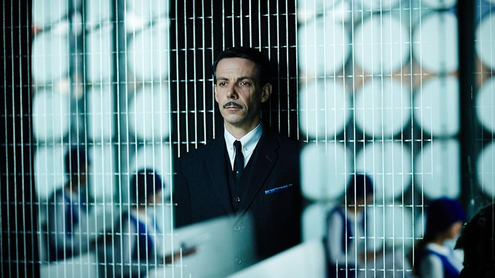 Ethan Hawke in a suit with mustache standing behind a mesh partition with uniformed people working at desks in the background
