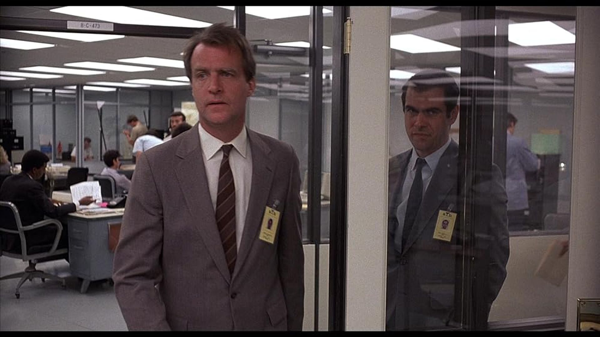 Two men in suits with passes in an office, one coming out of a glass door, the other standing behind it