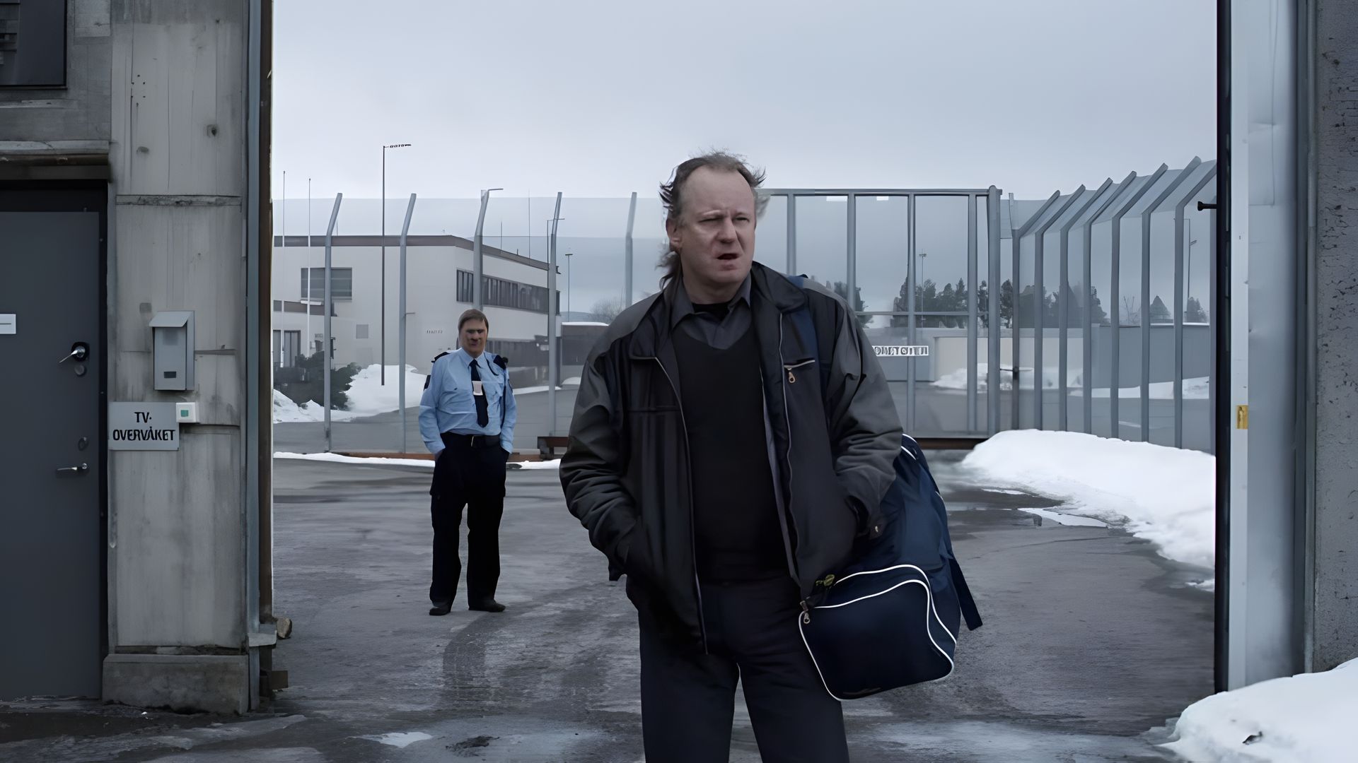 Stellan Skarsgård carrying a bag exits through prison gates, with a guard in the background, buildings and snow visible beyond the fence