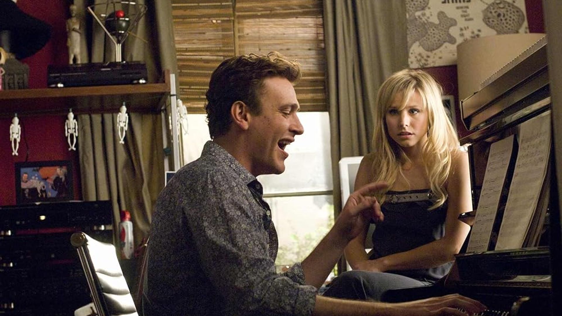 A man plays piano and sings while a woman nearby looks at him with bewilderment