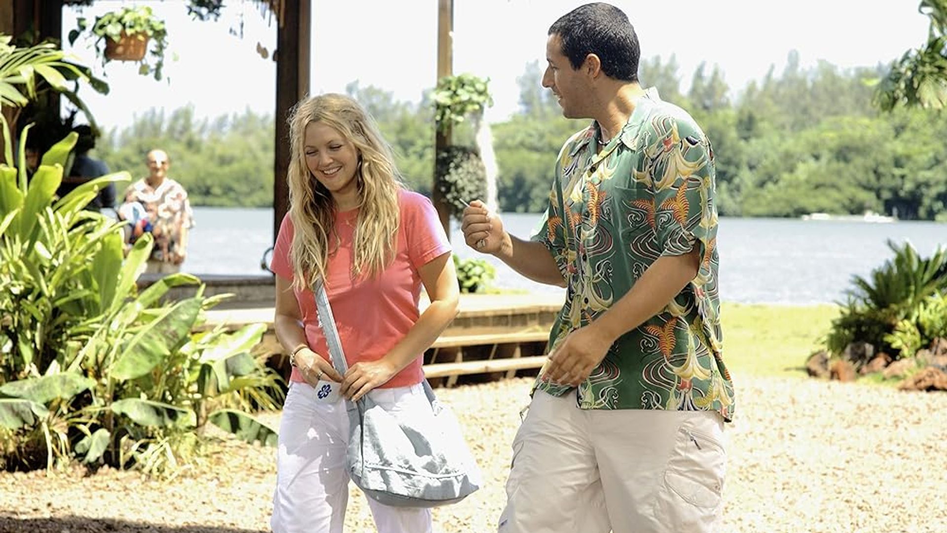 Drew Barrymore and Adam Sandler talking on a sunny beach with greenery and water in the background