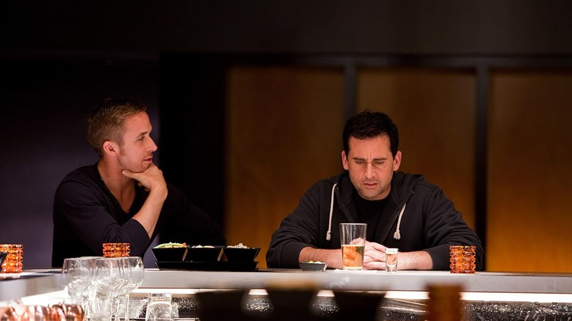 Ryan Gosling and Steve Carell sit at a bar counter, one gazing thoughtfully into his glass, the other watching
