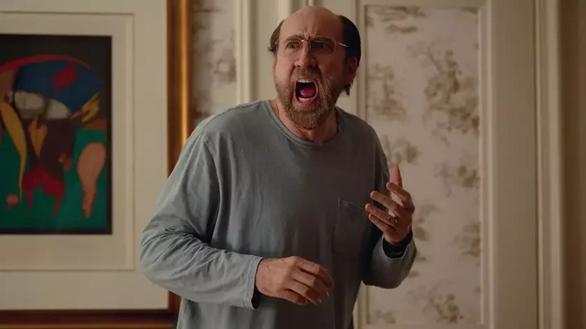 Nicolas Cage wearing glasses and a gray long-sleeved shirt screaming while standing in a room