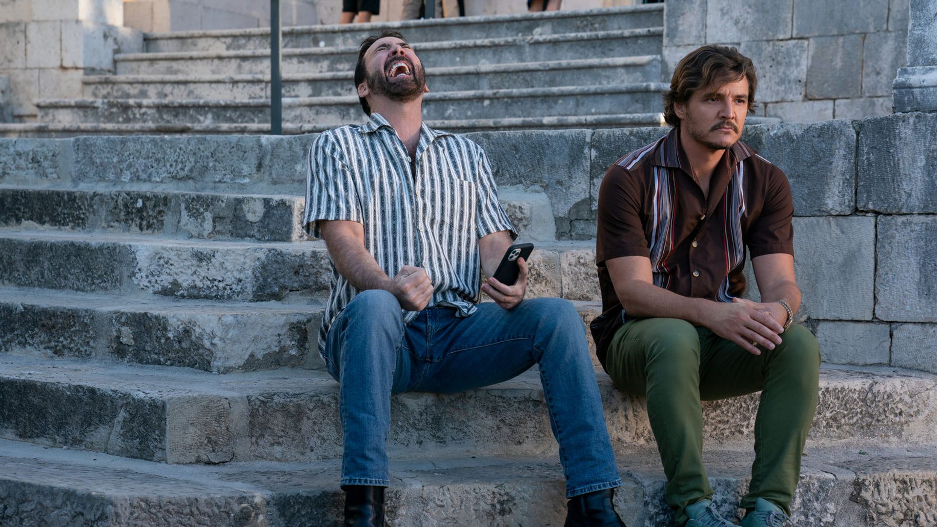 Nicolas Cage and Pedro Pascal sit on stone steps: one laughs with a phone in hand, the other gazes thoughtfully to the side