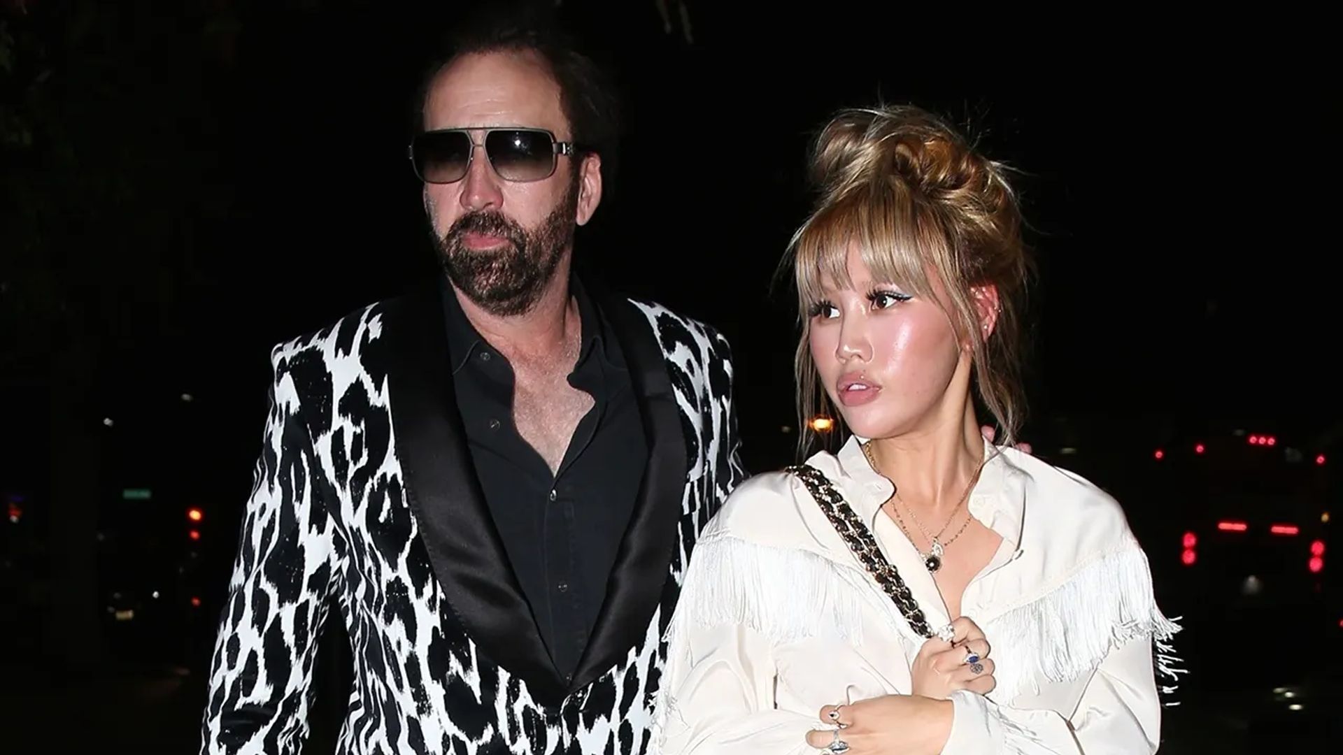 Nicolas Cage in a black and white patterned blazer and Erika Koike in a white blouse walking together at night
