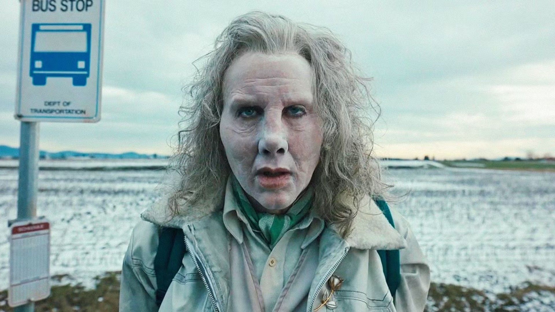 Nicolas Cage with gray hair and a pale face stands by a bus stop sign against a winter landscape backdrop