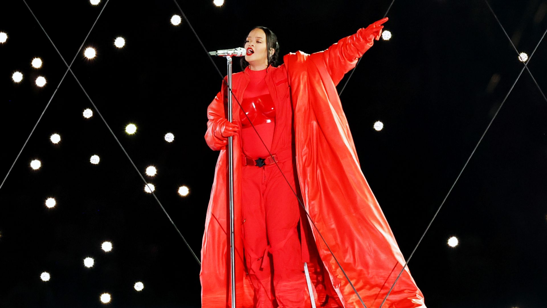 Rihanna in a bright red suit and long cape performs on stage with a microphone
