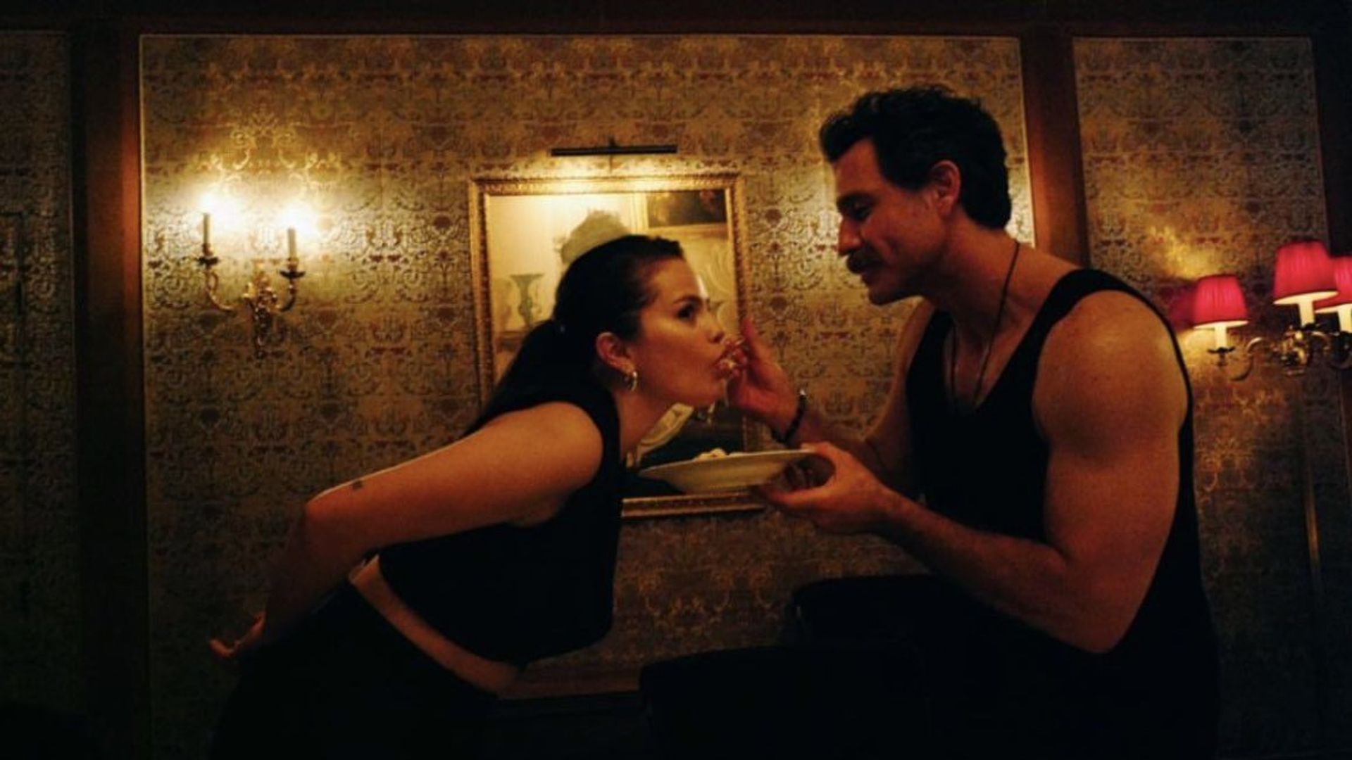 A man in a black tank top feeds Selena Gomez in the dim light of a room