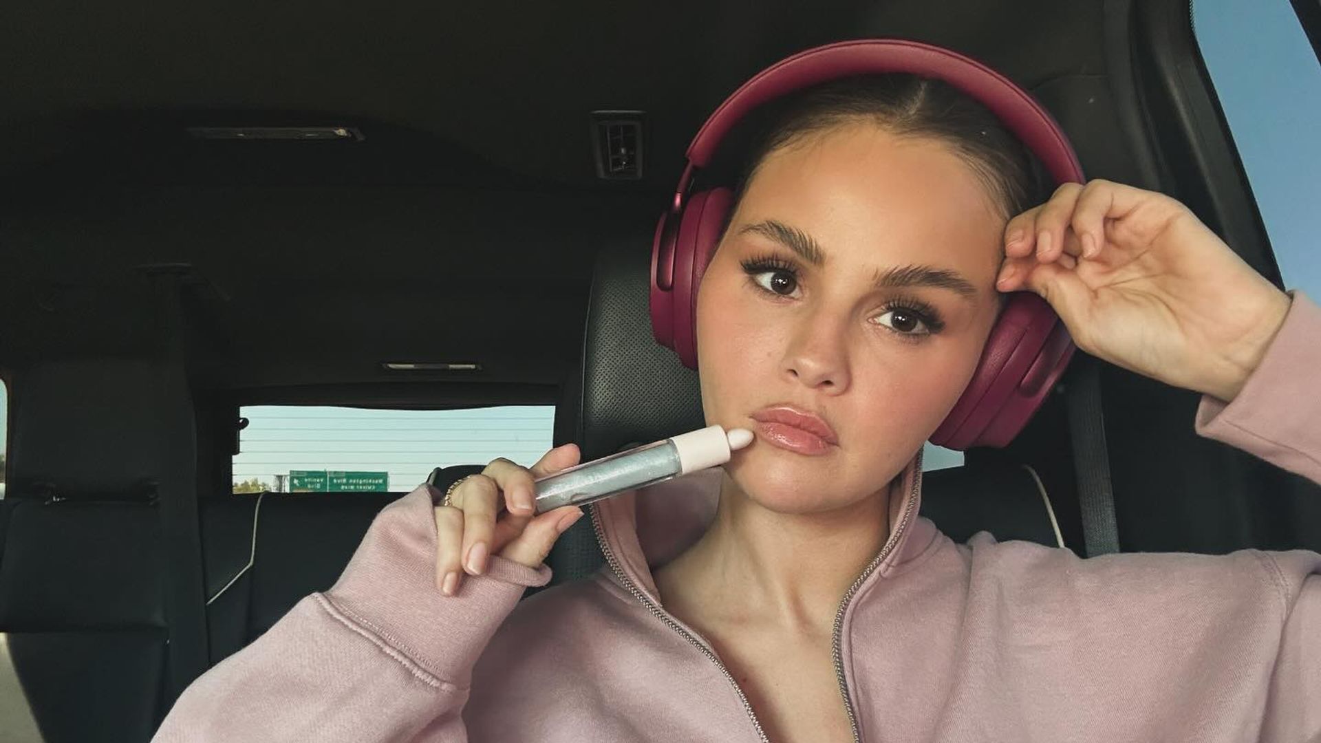 Selena Gomez in a pink sweatshirt and headphones sits in a car, holding a cosmetic product up to her face