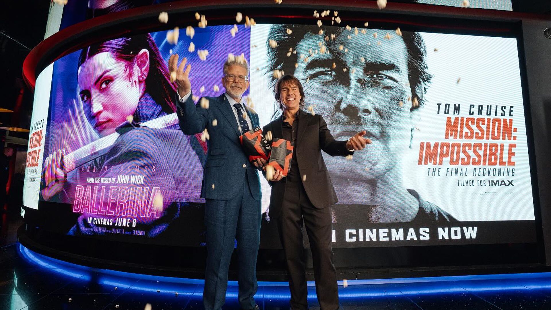 Two men in suits pose with popcorn boxes against a backdrop of movie posters for Ballerina and Mission: Impossible