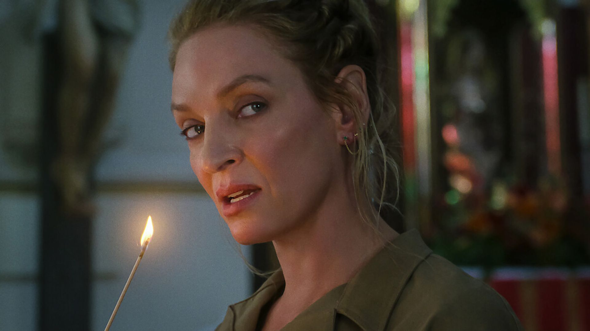 Uma Thurman with her hair pulled back holds a lit candle and looks to the side with a slightly blurred background