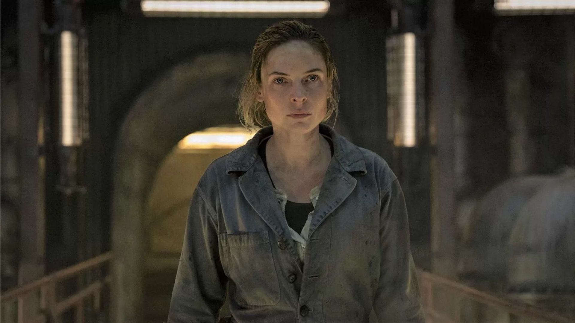 Rebecca Ferguson in work overalls stands in a dimly lit industrial space, looking straight ahead