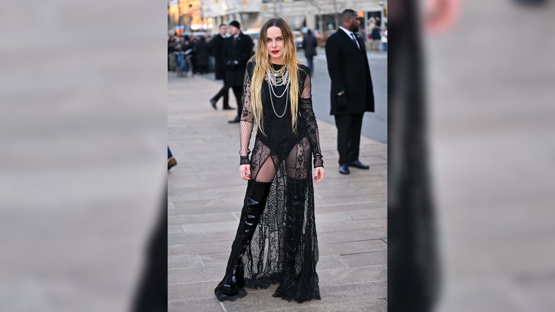 Rebecca Ferguson poses outdoors in a long sheer black dress with lace and high patent leather boots.
