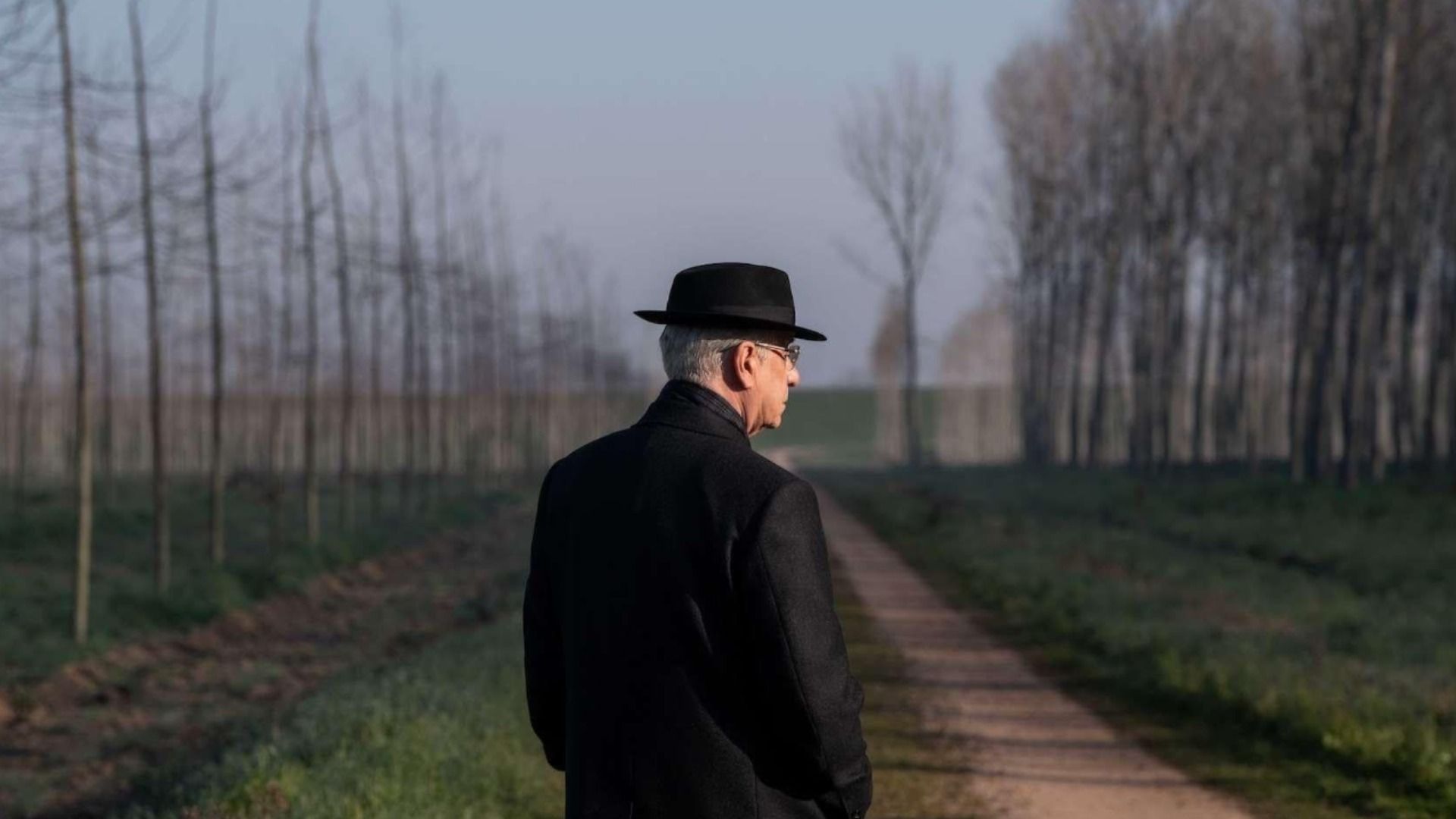 Actor Toni Servillo in a black coat and black hat stands next to trees