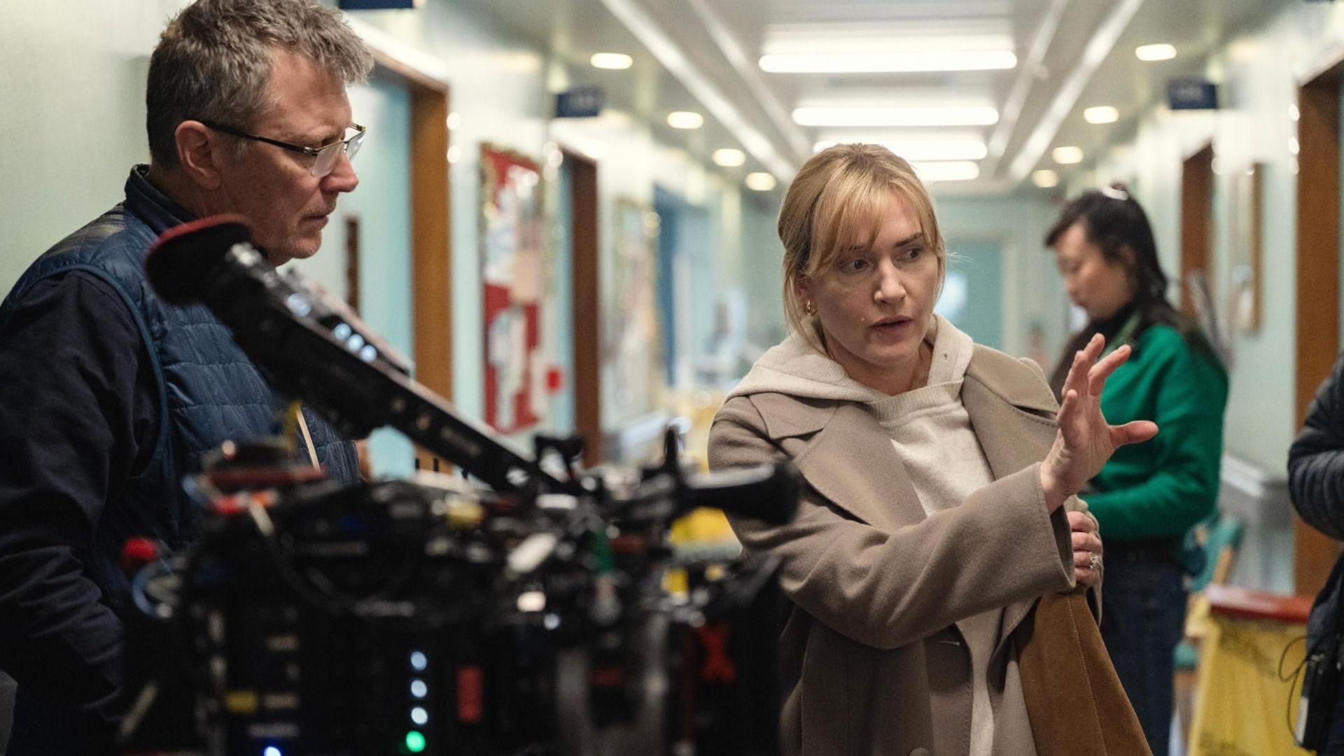 Actress Kate Winslet in a beige coat gives directions to a man with a camera