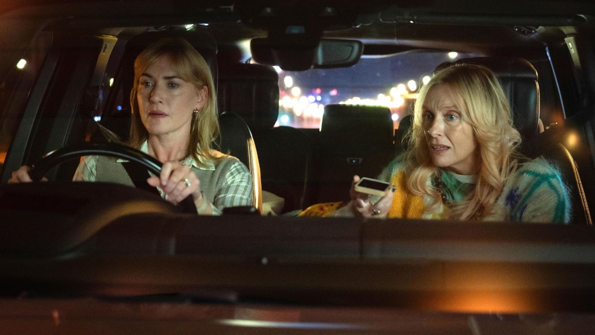 Actress Kate Winslet sits behind the wheel of a car while actress Toni Collette talks on the phone beside her