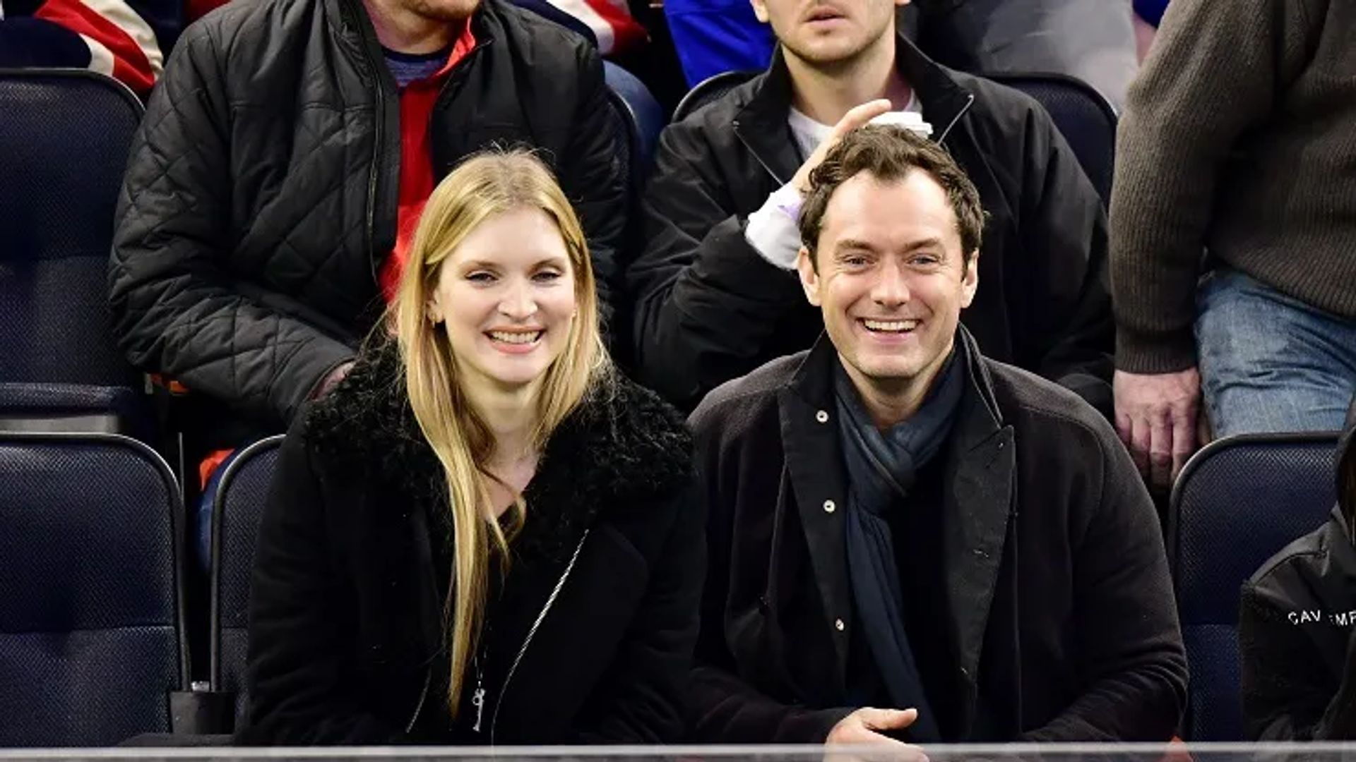 Jude Law and Phillipa Coan sit side by side in the stands, smiling and looking at the camera