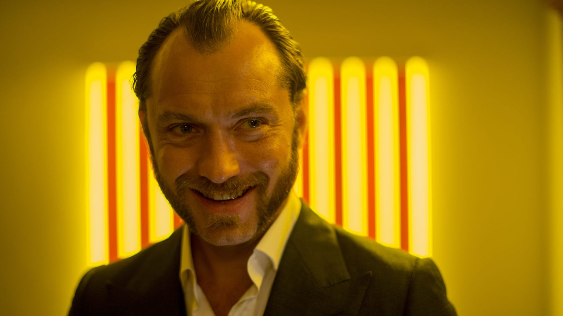 Jude Law in a suit jacket smiles against a backdrop of bright yellow light streaks