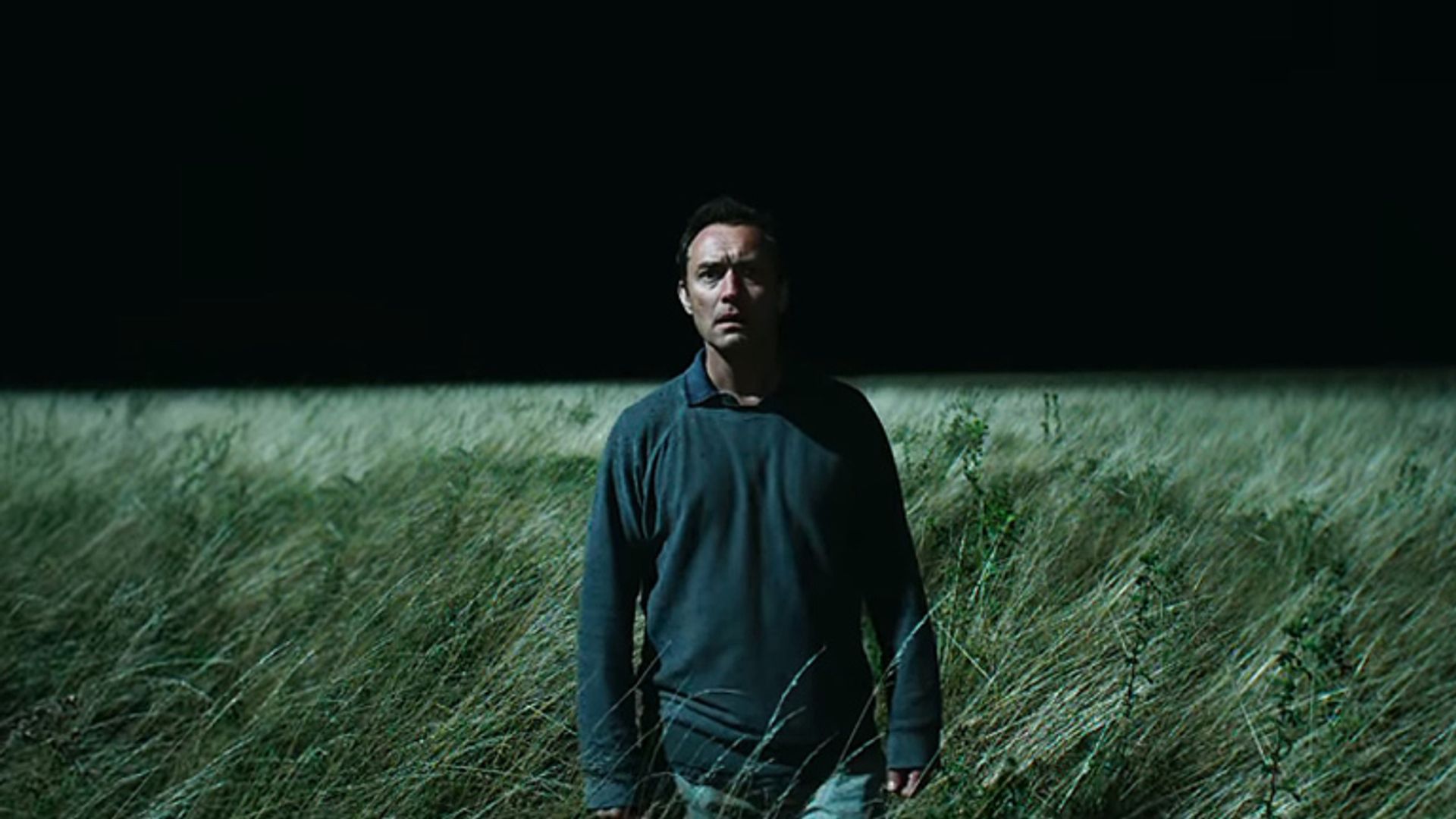 Jude Law in dark clothing stands in a tall grass field at night, illuminated by harsh light