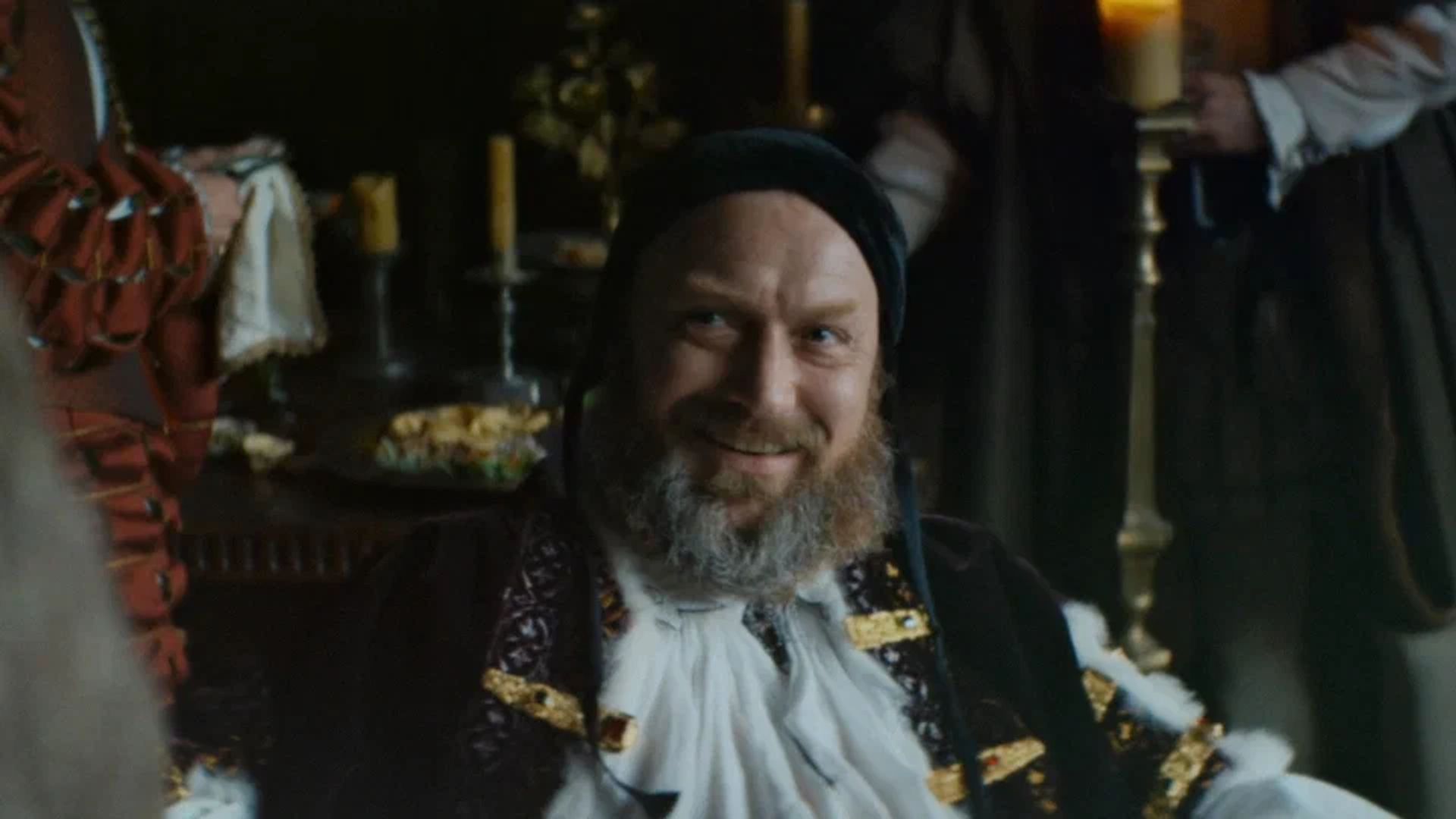 Jude Law in medieval costume with a beard smiles while sitting at a festive candlelit table