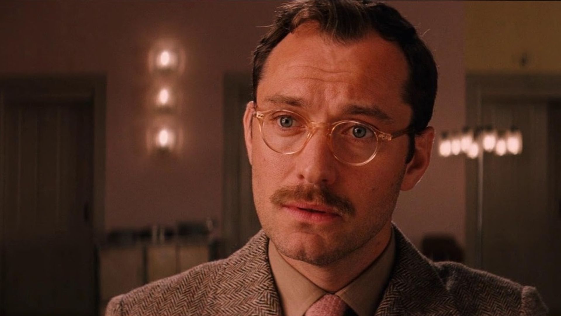 Jude Law wearing glasses and a mustache in a suit looks with a serious gaze in the warm interior of a room