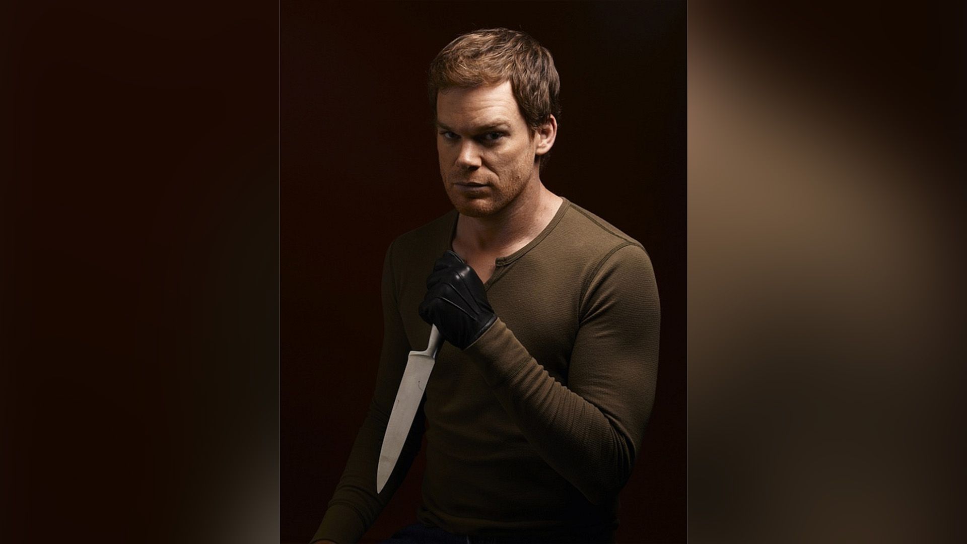 Michael C. Hall in a brown jacket and black gloves holding a knife in his hand