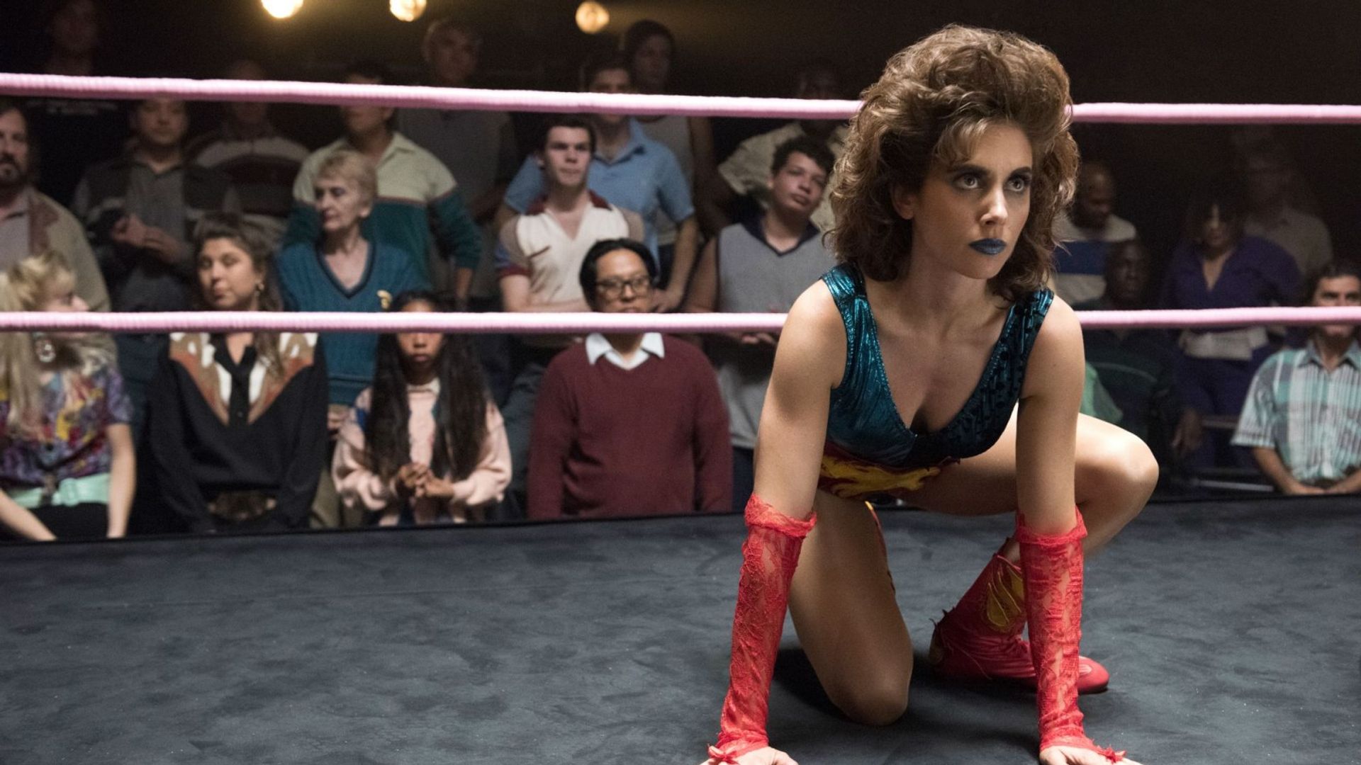 Alison Brie in wrestler costume sits in stance on the ring