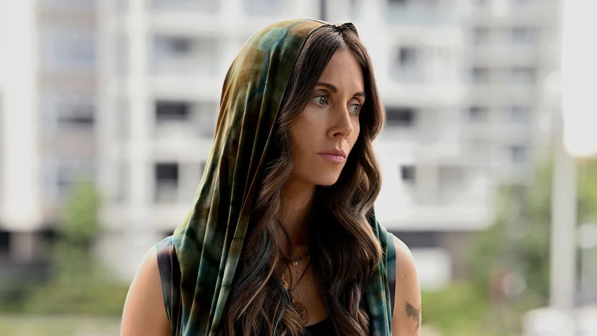 Alison Brie with long hair and a hood gazes thoughtfully to the side against a high-rise building backdrop