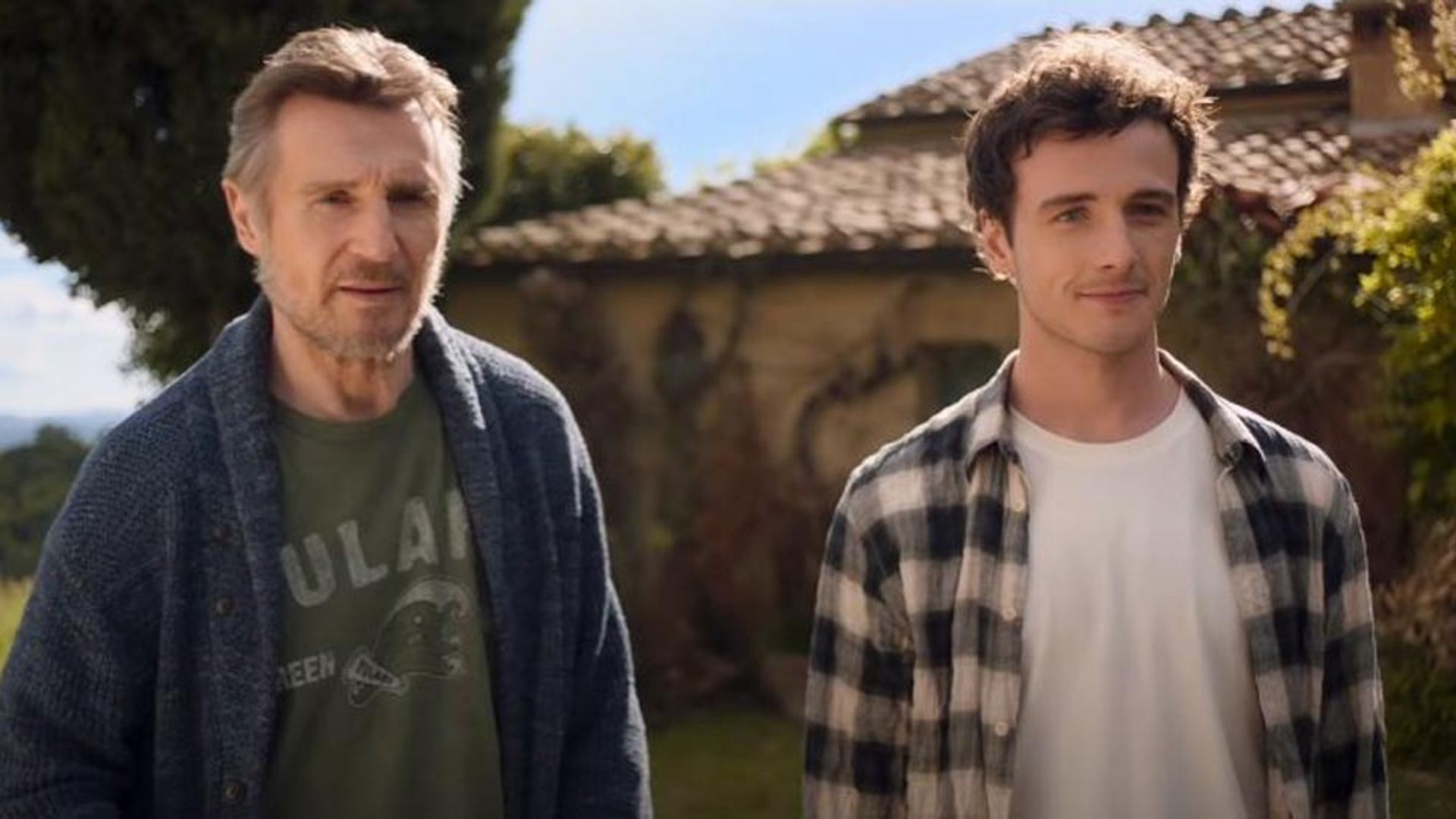 Liam Neeson and Michael Richardson stand side by side in front of a house with a tiled roof on a sunny day