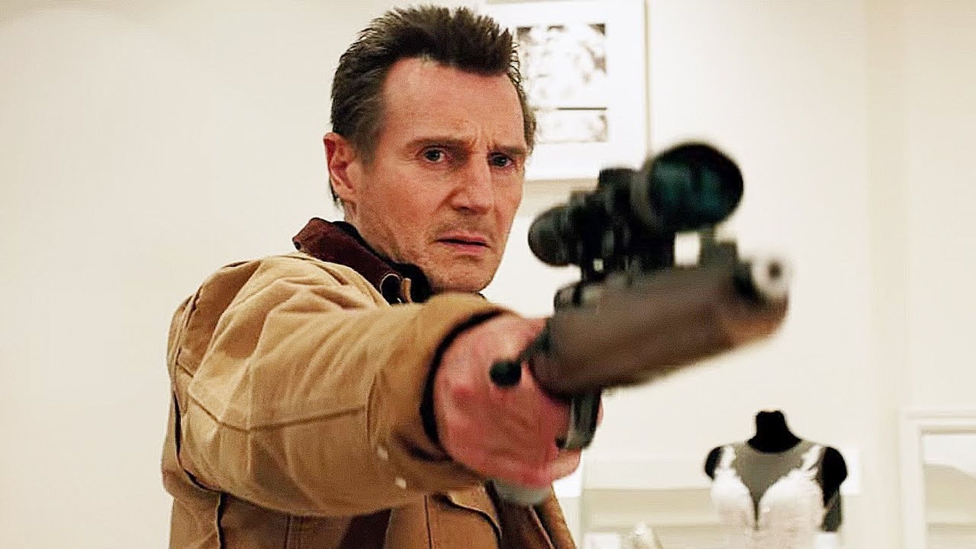 Liam Neeson in a beige jacket aims a scoped rifle, with a wedding dress mannequin behind him