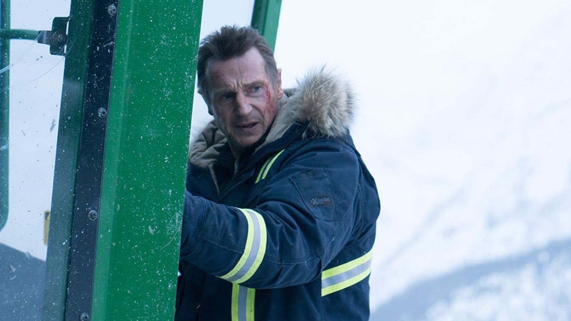 Liam Neeson in a hooded winter jacket grips the door of a green cabin with a snow-covered landscape in the background