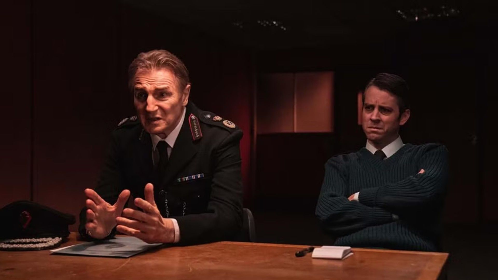 Liam Neeson in police uniform sitting at an interrogation table, scene from the series "Derry Girls"
