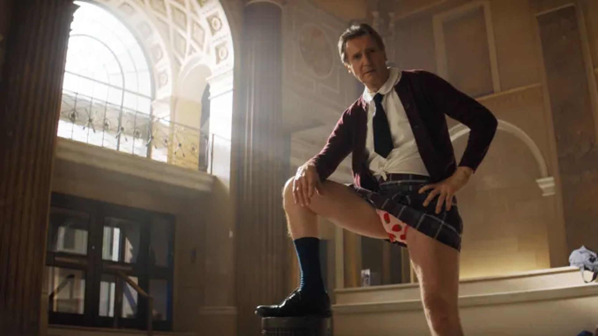 Liam Neeson in school uniform and tie poses with raised leg, exposing heart-patterned underwear, against a columned interior backdrop