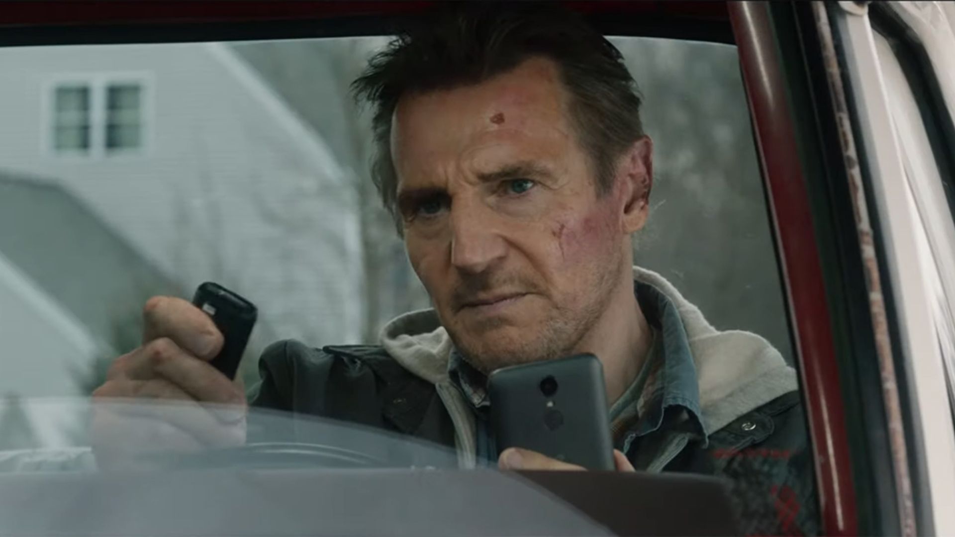 Liam Neeson sits in a car tensely staring at the phone and remote control in his hands