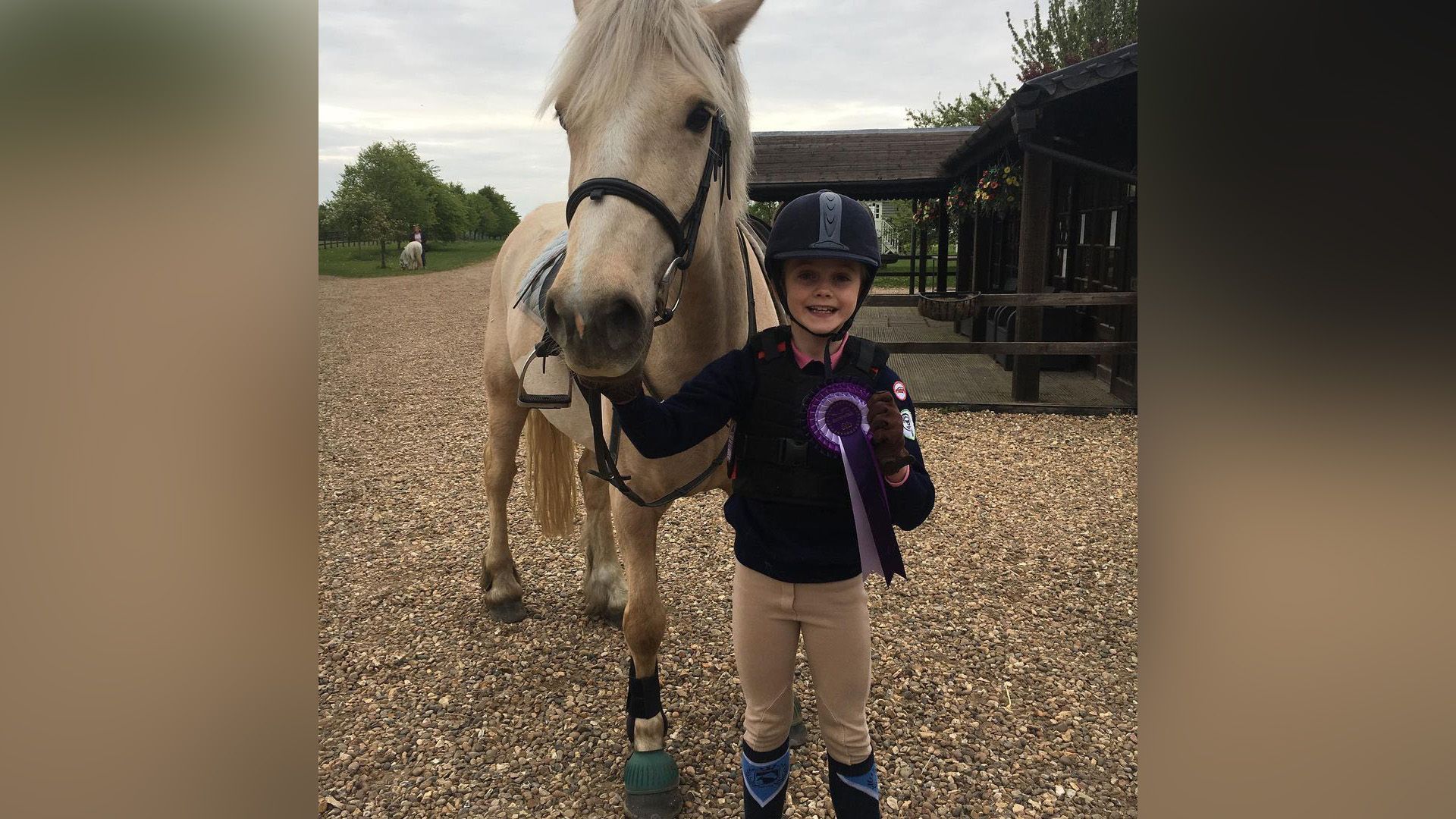 Evie Templeton as a child, dressed in riding attire and helmet, standing next to a horse while holding a purple award ribbon