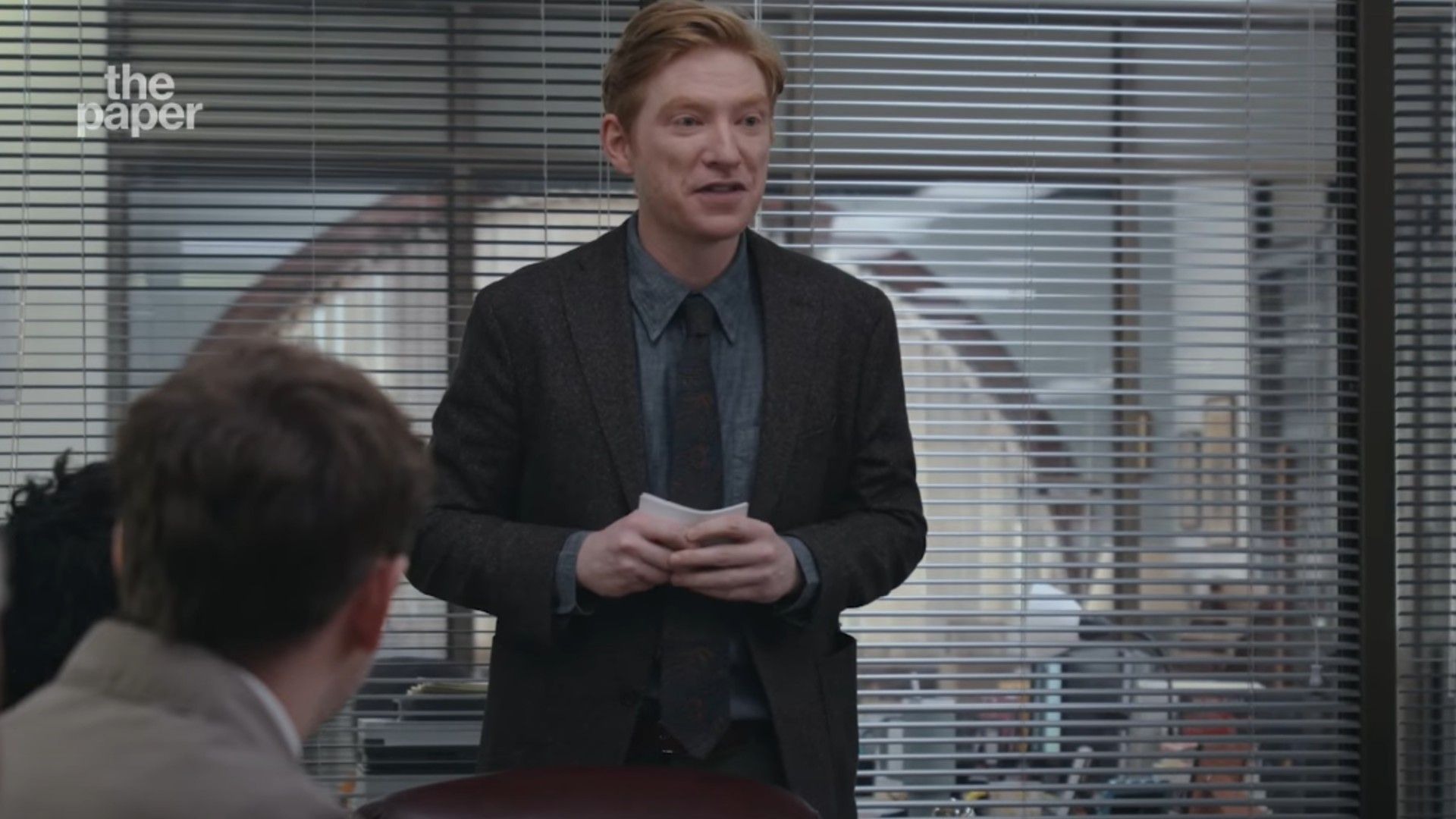 Domhnall Gleeson in a suit standing in a conference room