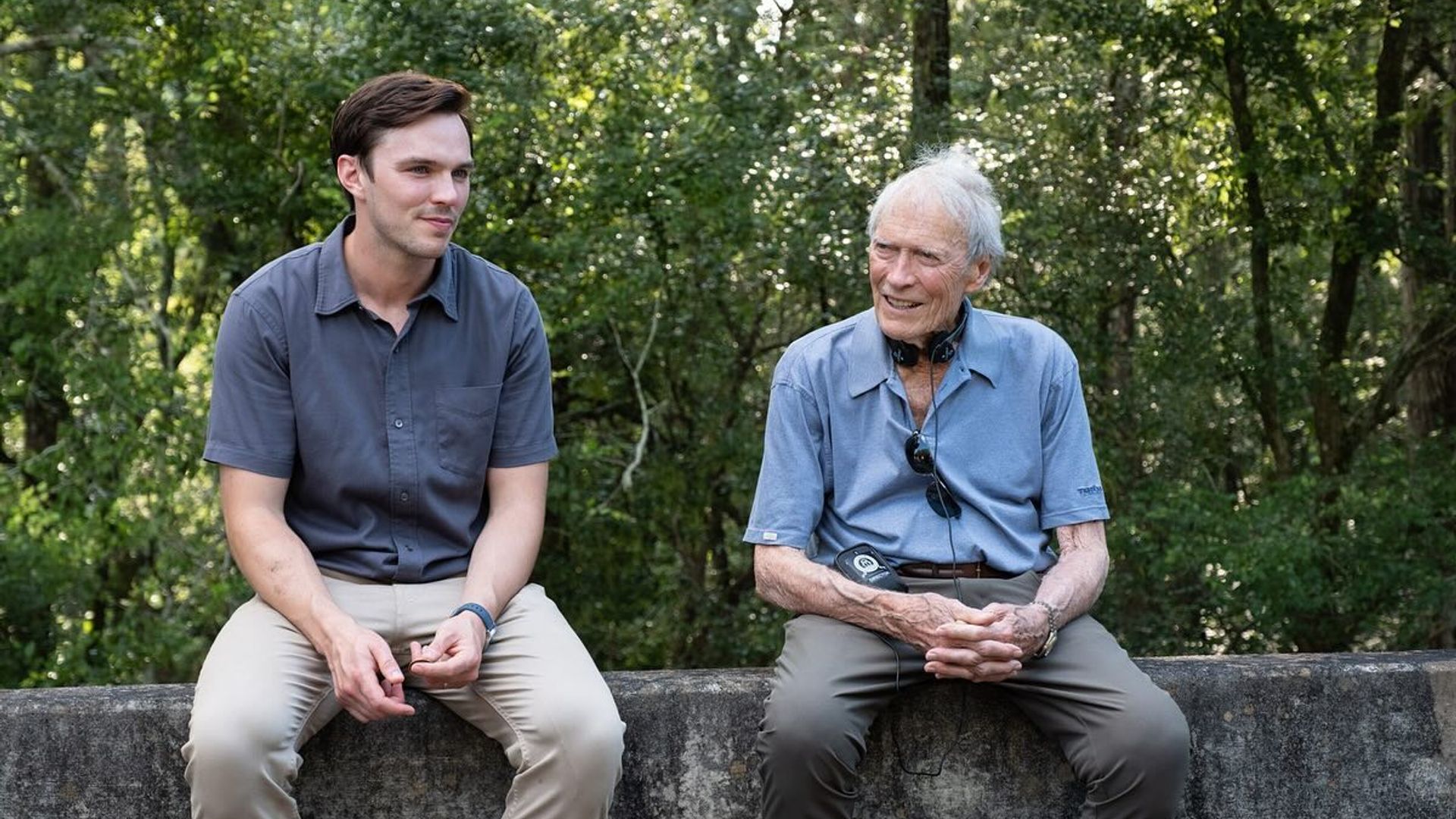 Nicholas Hoult and Clint Eastwood in casual clothes sitting on a concrete barrier and talking with a forest backdrop