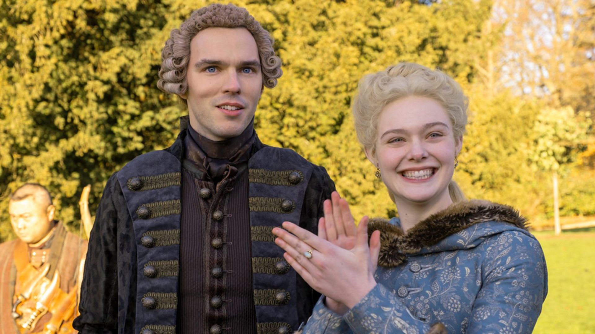 Nicholas Hoult and Elle Fanning in 18th-century period costumes standing on the street, the actress is smiling and clapping her hands