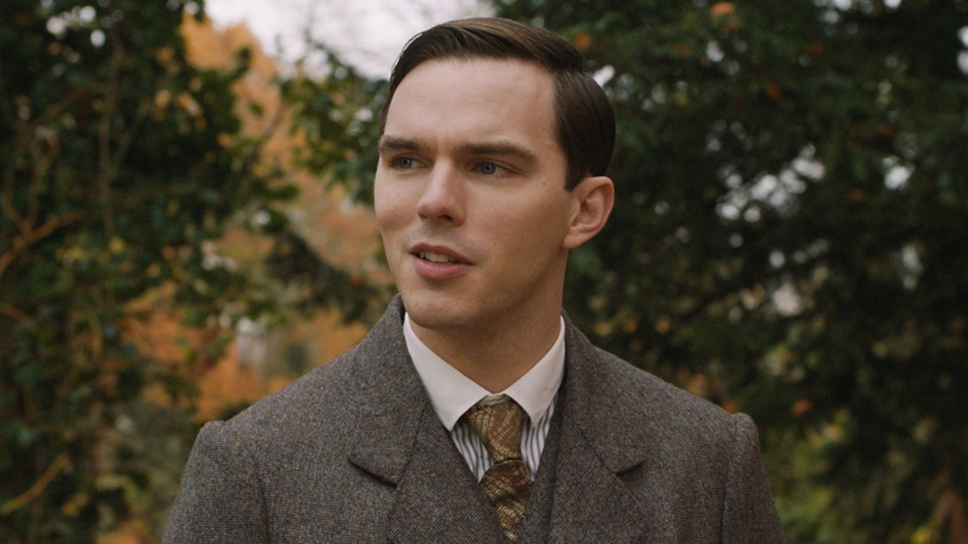 Nicholas Hoult in a brown suit and tie standing on the street in the role of young J.R.R. Tolkien