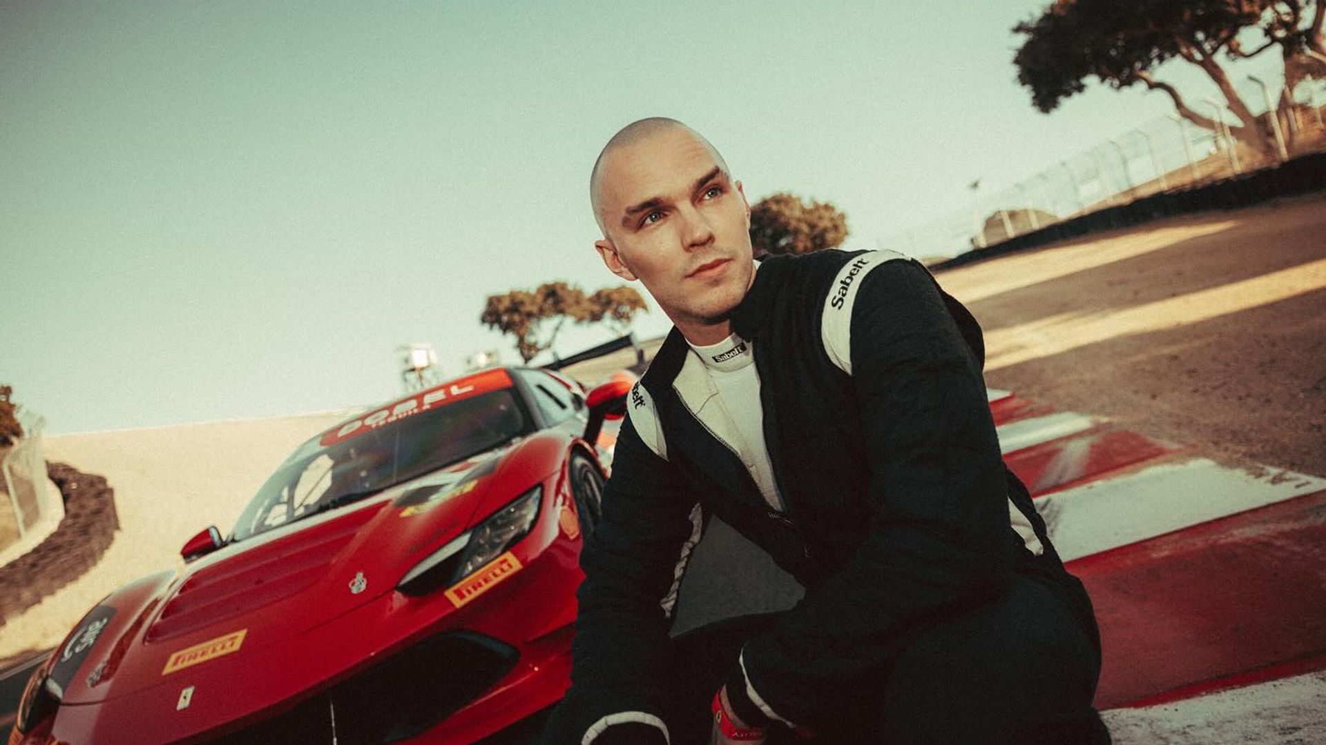 Nicholas Hoult in a racing suit sits on the track with a red Ferrari sports car behind him
