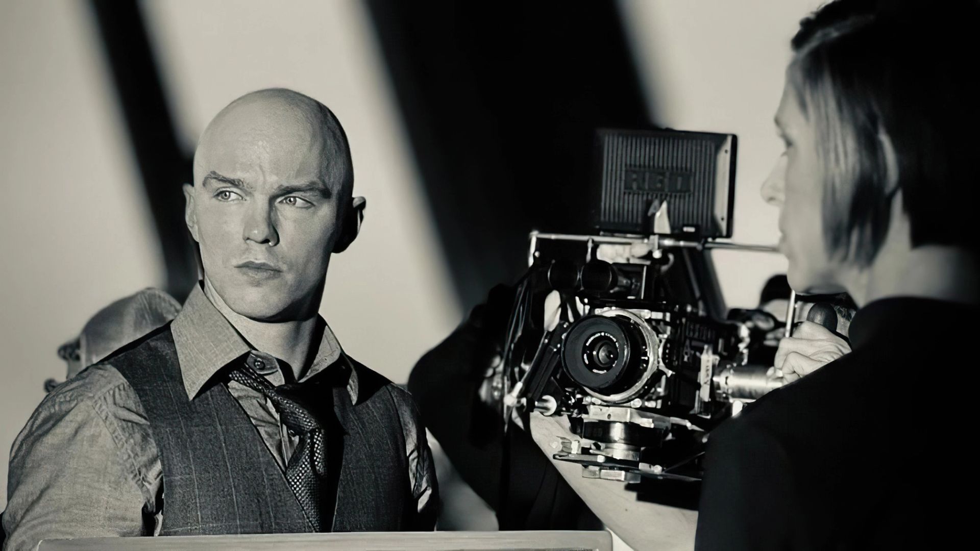 Nicholas Hoult in a vest and tie stands next to a professional film camera on a movie set